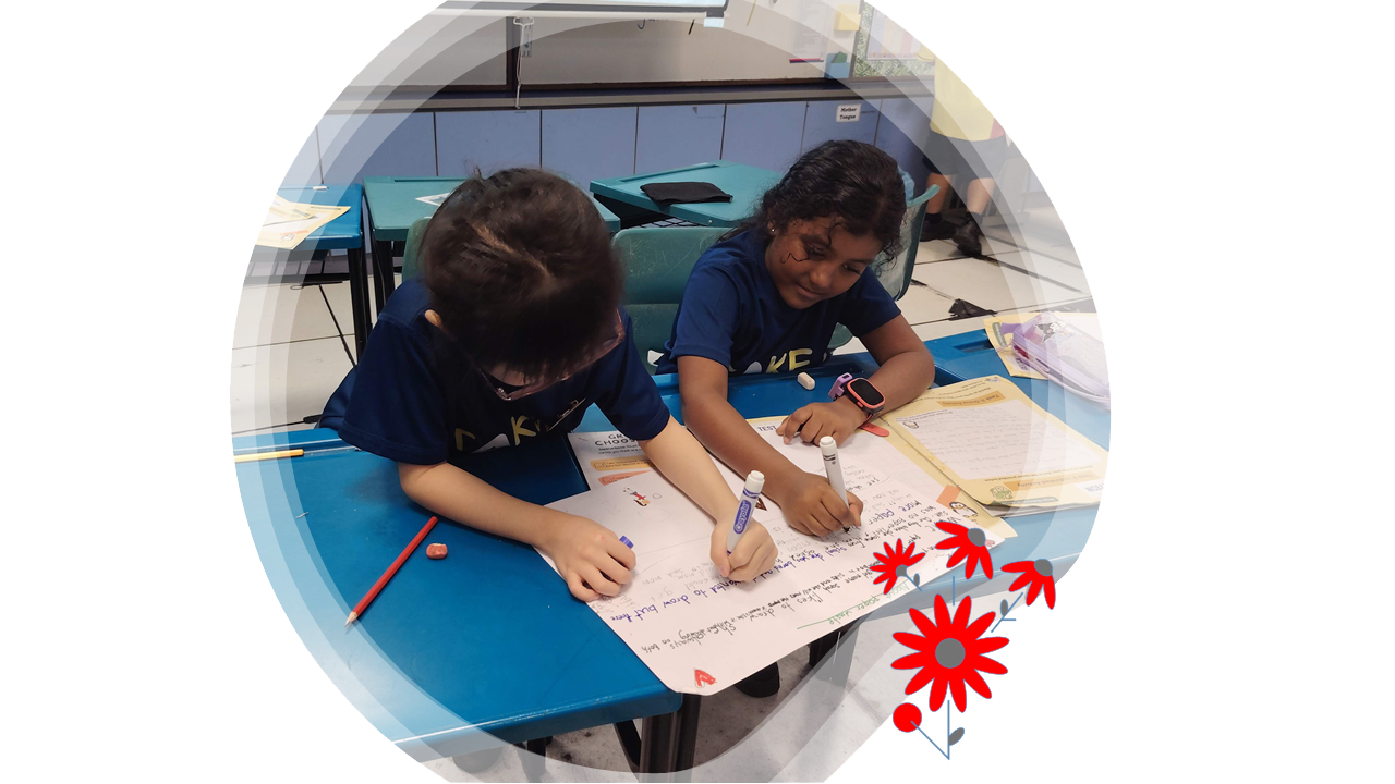 Two students sit side by side at a classroom desk, writing and drawing together on a large sheet of paper.